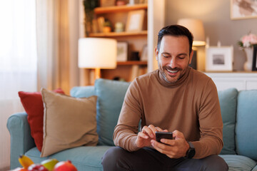 Obraz premium Portrait of handsome man smiling and scrolling apps over smart phone while sitting on sofa at home