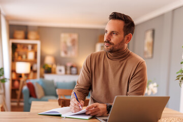 Serious businessman looking away thoughtfully while writing notes and working over laptop at desk