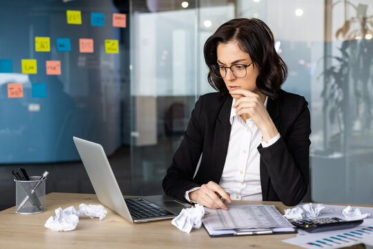 Businesswoman in office intensely analyzing financial reports on laptop with scattered crumpled paperwork around. Represents focus, analysis, and problem-solving in modern business environment.
