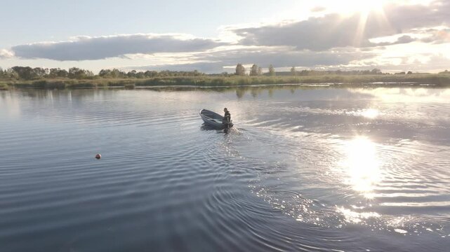 A person rows a small boat across a wide lake. Sunlight reflects on the water, with clouds in the sky. Trees line the horizon, and a buoy floats nearby.