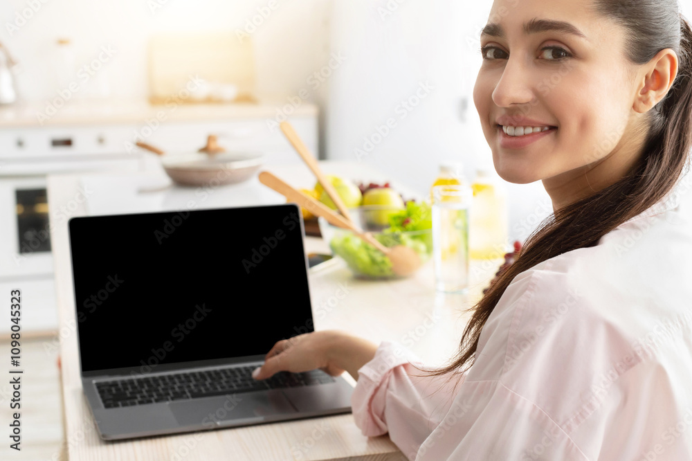 Aufkleber Portrait of happy smiling young lady using personal computer with black empty screen for mock up design, looking back sitting at dinner table in kitchen, working and typing on keyboard