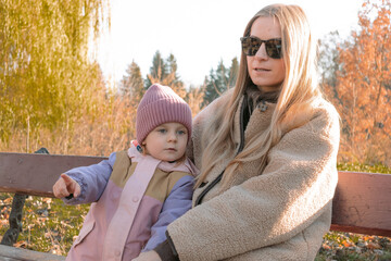 Fototapeta premium A happy girl and a young mother are sitting on a bench in autumn on a walk in nature