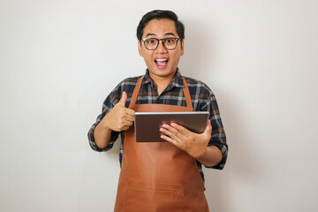 Asian man wearing apron holding tablet and showing thumbs up gesture with happy face, coffee shop...