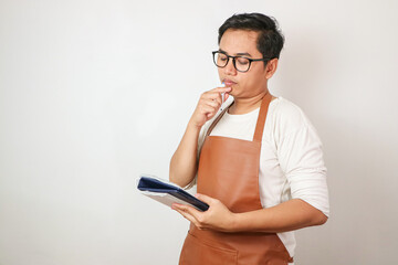 Asian barista man in brown apron taking order, writing on menu book list, thinking with pen on head. Isolated on white background. - Powered by Adobe