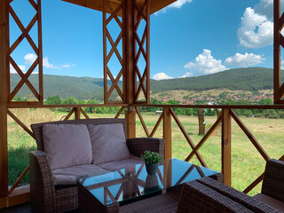 A cozy outdoor seating area with a wicker sofa and glass table, overlooks a scenic landscape of green hills and a distant village under a bright blue sky with fluffy clouds
