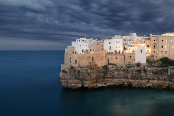 Ancient mediterranean town on rocky seaside against cloudy sky after sunset, Polignano a Mare, Italy