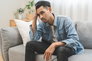 Fototapeta premium Sad unhappy, alone asian young man headache holding forehead thinking about problem, difficulty, feeling failure and exhausted, suffering sitting on sofa, grief sorrow and bad relationship or break up