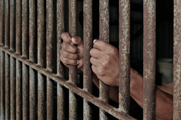 hands of a prisoner behind prison bars on black background	
