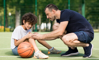Side view portrait of focused adult man coach helping boy with knee trauma after playing basketball...