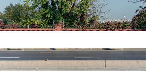 Large blank billboard on a brick wall alongside a road, with trees and a cityscape in the background, ideal for advertising mockups © Bonsales