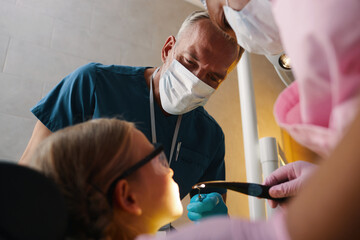 Dentist wearing mask examining a child's teeth while dental assistant holding tools. Child sitting...