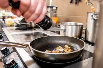 Close-up of a hand using a pepper mill while cooking in a professional kitchen. A frying pan sits...