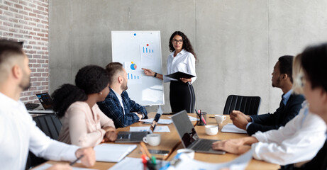 Fototapeta premium Latin Businesswoman Making Business Presentation Giving Speech For Colleagues Standing And Pointing At Blackboard With Charts And Graphs In Modern Office. Panorama, Selective Focus
