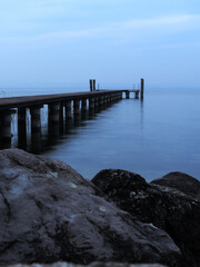 Obraz premium Muelle de madera en la costa de un lago con rocas, en un día nublado