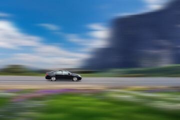 Black car in motion, driving along a road with a blurred background of greenery, a blue sky with white clouds, and distant mountains. Fast moving sedan with motion blur effect