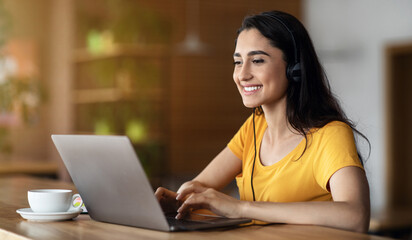 Joyful girl student studying online at cafe, using laptop and earphones, empty space