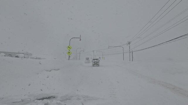 SLOW MOTION: Driving carefully through road curve during a heavy snowstorm with vehicles cautiously navigating. Visibility is reduced, traffic signs and poles are covered with thick layers of snow.