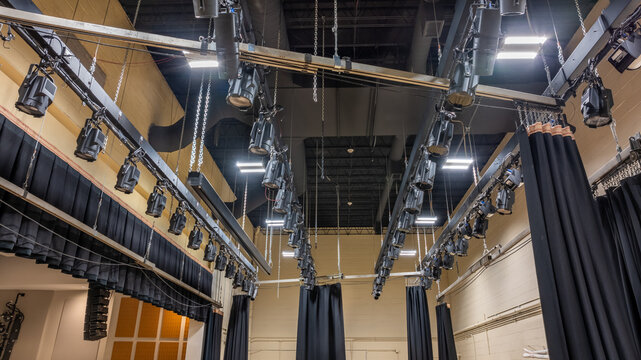 Theater stage rigging with theatrical equipment, lighting, supports, and chains. Photo taken from an elevated position looking across the rigging.	