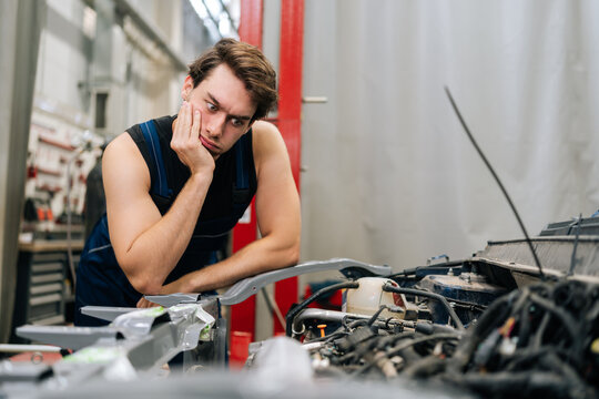 Disappointed automotive mechanic male in grease-stained uniform, leans against car, contemplates fixing broken engine in workshop bustling with tools and parts. Concept of car repair and maintenance.