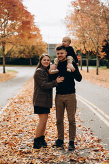Fototapeta premium A family portrait with a baby on the father's shoulders. The young couple shows warm emotions, the mother hugs the father while he holds the child. Autumn alley with fallen leaves.