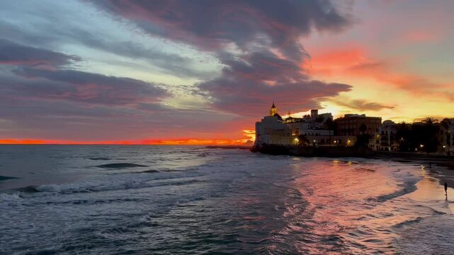 Video del atardecer pueblo Sitges, Barcelona. La luz del sol ha te&ntilde;ido las nubes de naranja creando reflejos en las aguas del mar y en la arena de la orilla, una estampa inolvidable, fondo de pantalla
