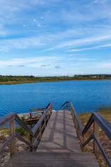 A wooden boardwalk with railings gently descends towards a calm lake, inviting visitors to approach...