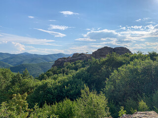 A lone figure perches atop a rocky outcrop, overlooking the vast expanse of the Rhodope Mountains under a clear blue sky.
