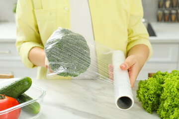Woman putting plastic food wrap over broccoli at countertop in kitchen, closeup