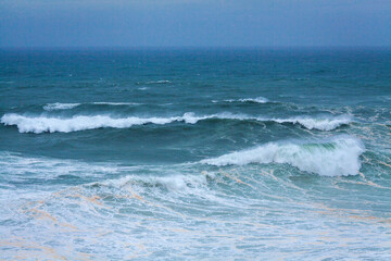 A vast, powerful ocean with rolling waves and white foam stretches under a soft, misty blue sky, creating a serene yet dynamic seascape