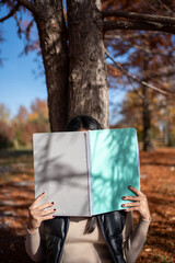 Student hiding face with notebook in autumn park
