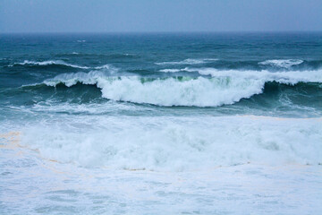 A vast, powerful ocean with rolling waves and white foam stretches under a soft, misty blue sky, creating a serene yet dynamic seascape