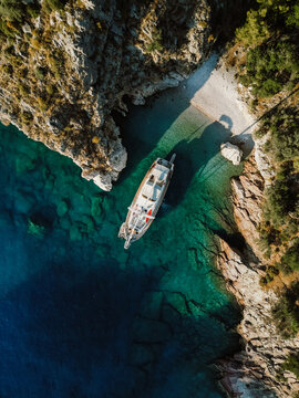 A stunning aerial view of a secluded cove with turquoise waters, rugged cliffs, and a luxury sailing yacht anchored near a small, hidden beach. A serene and picturesque escape in nature in T&uuml;rkiye