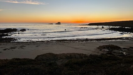 A stunning coastal scene at sunset, featuring a sandy beach with rocky outcrops and gentle waves. The sky transitions from warm oranges to deep blues, creating a peaceful and picturesque view.