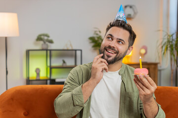Happy man wears festive birthday hat holding cupcake makes wish joyful congratulating blowing...