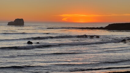 A beautiful sunset over a rocky shoreline with gentle waves and an isolated rock formation in the distance, casting a warm orange glow across the sky and water. - Powered by Adobe