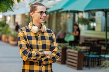 Obraz premium Portrait of happy man with headphones around neck smiling friendly, glad expression looking at side dreaming, resting, relaxation feel satisfied good news outdoors. Young guy in urban city street.