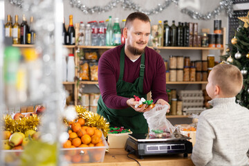Caring seller weighing candies for smiling little customer in market with Xmas decoration