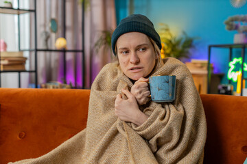 Young woman covered by warm blanket drinks coffee at home. Caucasian female freezes from the cold,...