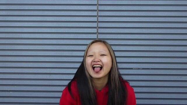 cheerful young asian girl in red hoodie teasing and making grimaces against city wall background, korean woman making fun and showing tongue at camera outdoors