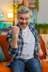 Obraz premium Happy senior man watching TV shouting, celebrating success, winning, goal achievement good news, lottery luck, victory sitting on sofa in living room. Mature old guy clenching fists at home. Vertical