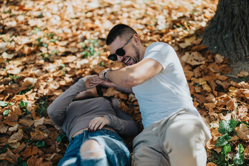 A young couple laughing and playing together on fallen autumn leaves in a sunny park. Their joyful expressions and casual attire convey a sense of happiness and carefree moments.