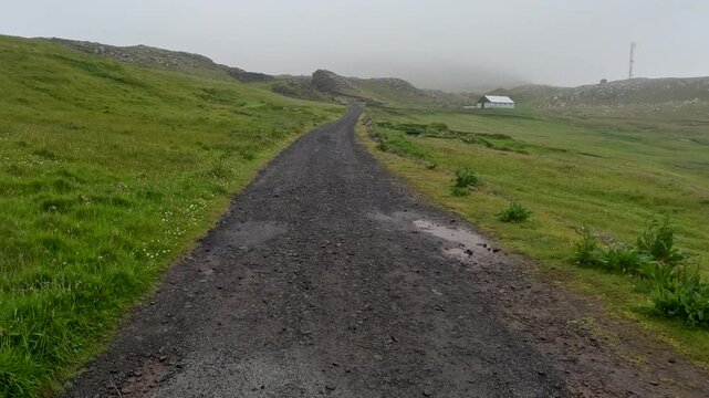 Moving backwards along a long gravel road or trail running through a mountain. Fog and rain with low cloud cover. Located on the remote Scandinavian island of N&oacute;lsoy within the Faroe Islands.