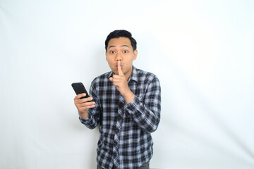 Fototapeta premium asian man holding mobile phone showing silent gesture with finger on lips isolated on white