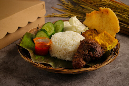 Delicious plate of Indonesian food featuring ayam bakar (roasted chicken), served with a mound of white rice, crispy cracker, tempeh, and fresh vegetables like cucumber and tomato on a woven basket