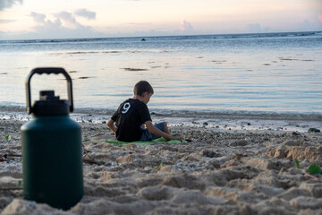 Boy sits on the beach with green towel and water bottle during sunset