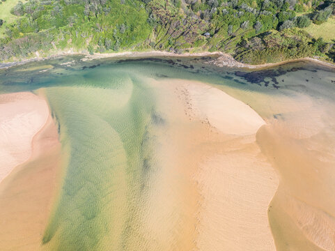 Aerial view of sandbars choking the exit of a narrow creek flowing near the base of green forest