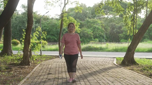 A middle-aged, overweight Asian woman walking for exercise on a park road in the evening, dressed in athletic wear and holding a water bottle, symbolizing a commitment to a healthy lifestyle.