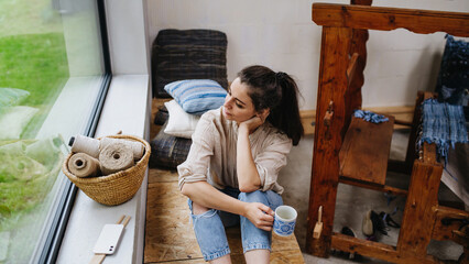 Young businesswoman taking break, sitting next to wooden loom and drinking coffee. Small weaving...