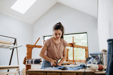 Young businesswoman working on workbench, preparing old fabric for weaving. Upcycled textile, small weaving business and circular economy.