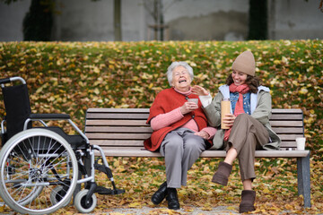 Portrait of a granddaughter and grandmother sitting on bench in autumn park, drinking coffee and...